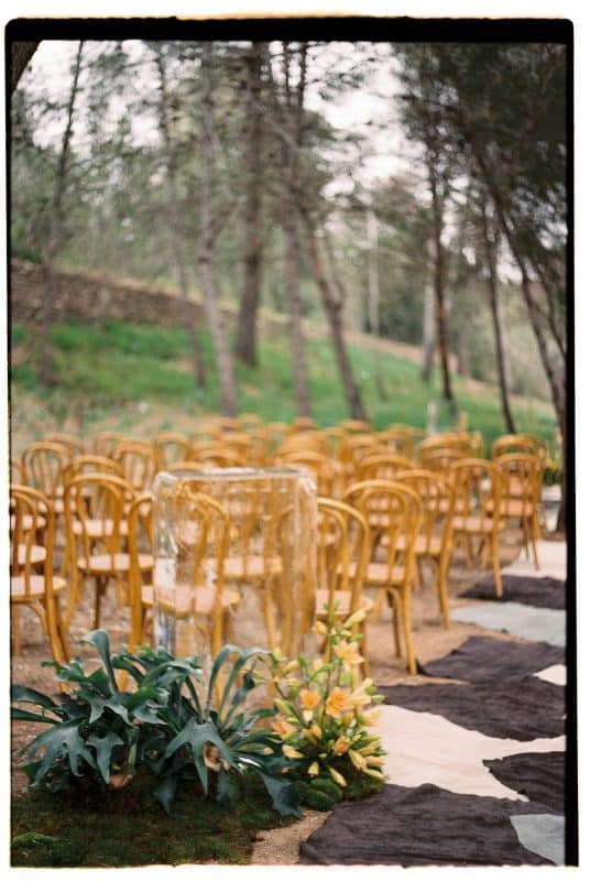 Pareja de novios en ceremonia civil al aire libre con sillas madera, flores frescas y decoración natural entre árboles.