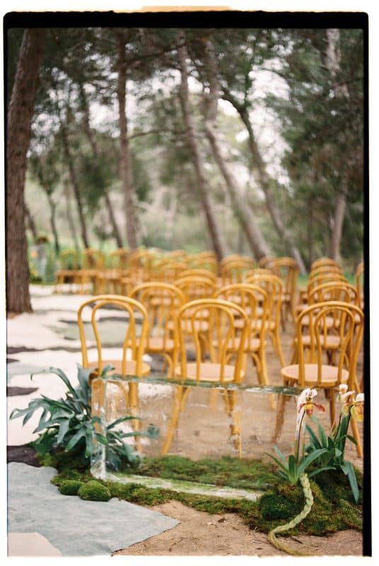 Pareja de novios en ceremonia civil al aire libre con sillas madera, flores frescas y decoración natural entre árboles.