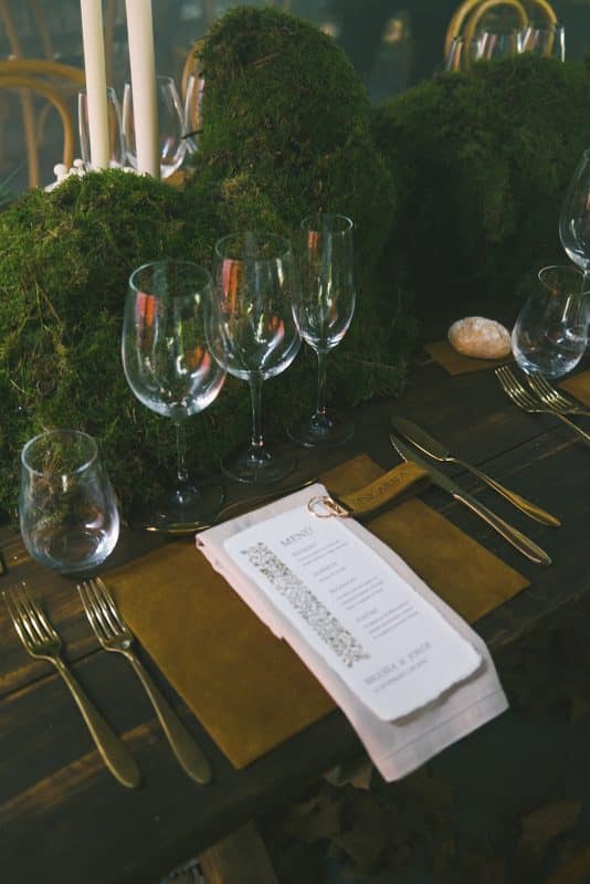 Mesa larga decorada con velas altas, musgo y cristalería en un ambiente estilo bosque encantado para cena de boda elegante.