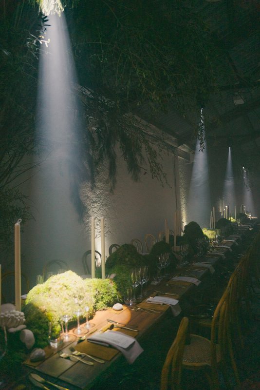 Mesa larga decorada con velas altas, musgo y cristalería en un ambiente estilo bosque encantado para cena de boda elegante.