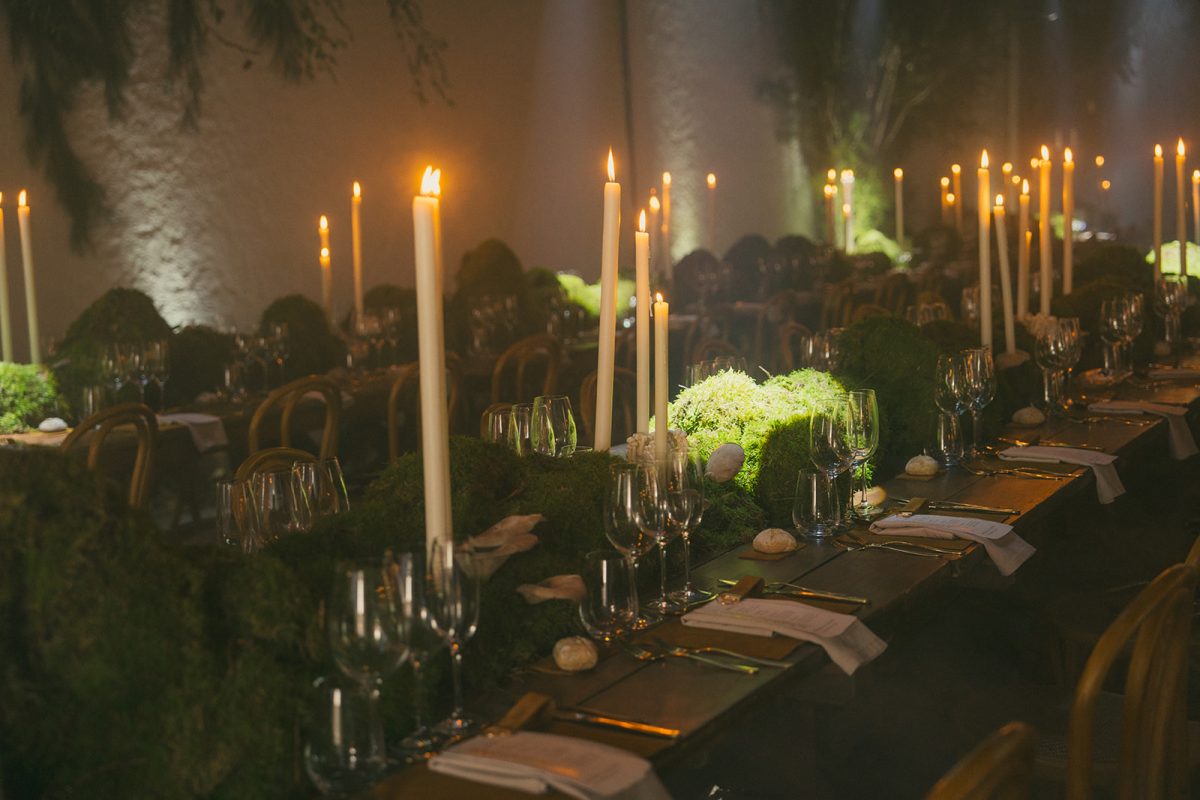 Mesa larga decorada con velas altas, musgo y cristalería en un ambiente estilo bosque encantado para cena de boda elegante.