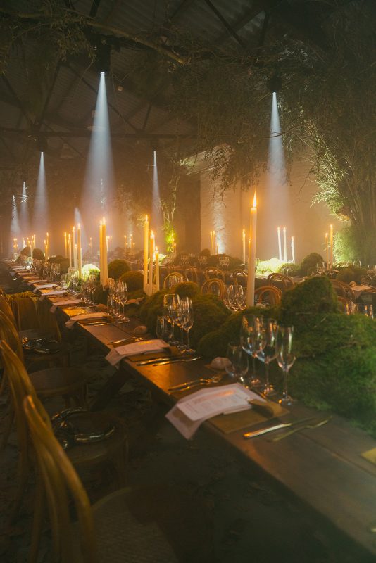 Mesa larga decorada con velas altas, musgo y cristalería en un ambiente estilo bosque encantado para cena de boda elegante.