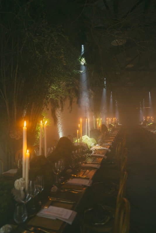 Mesa larga decorada con velas altas, musgo y cristalería en un ambiente estilo bosque encantado para cena de boda elegante.
