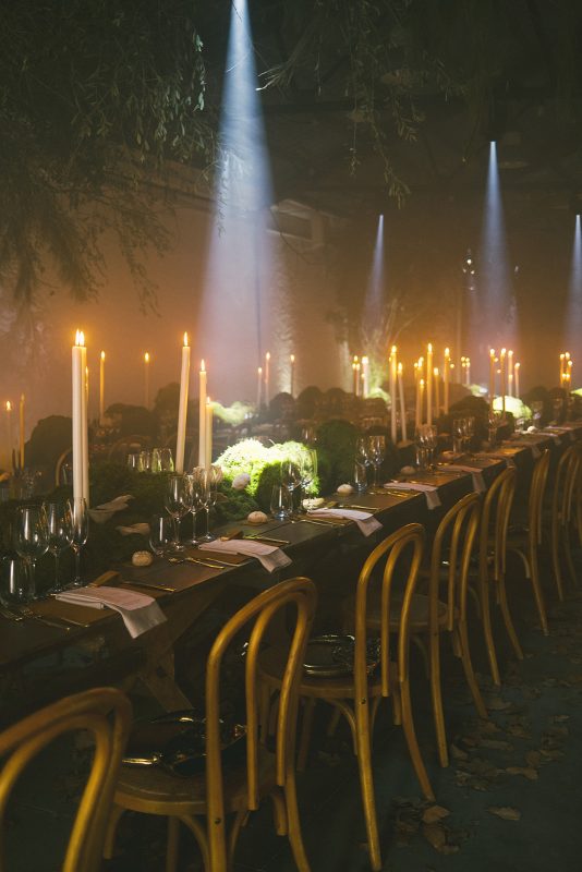 Mesa larga decorada con velas altas, musgo y cristalería en un ambiente estilo bosque encantado para cena de boda elegante.