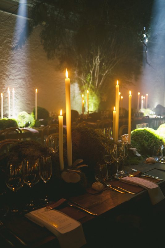 Mesa larga decorada con velas altas, musgo y cristalería en un ambiente estilo bosque encantado para cena de boda elegante.