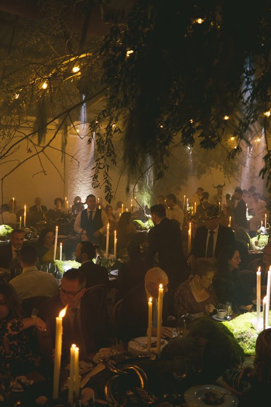 Mesa larga decorada con velas altas, musgo y cristalería en un ambiente estilo bosque encantado para cena de boda elegante.