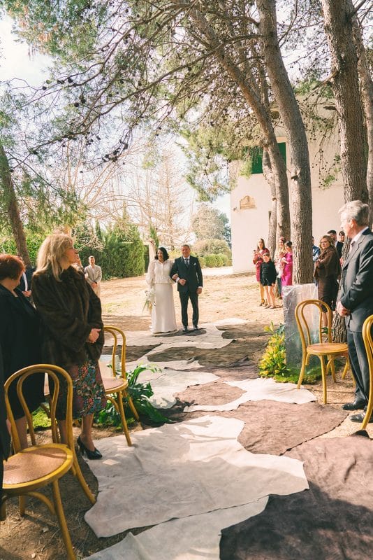 Pareja de novios en ceremonia civil al aire libre con sillas madera, flores frescas y decoración natural entre árboles.