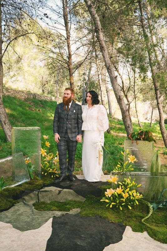 Pareja de novios en ceremonia civil al aire libre con sillas madera, flores frescas y decoración natural entre árboles.
