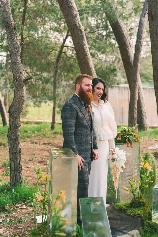 Pareja de novios en ceremonia civil al aire libre con sillas madera, flores frescas y decoración natural entre árboles.