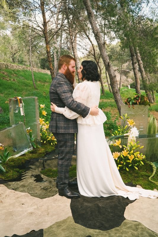Pareja de novios en ceremonia civil al aire libre con sillas madera, flores frescas y decoración natural entre árboles.