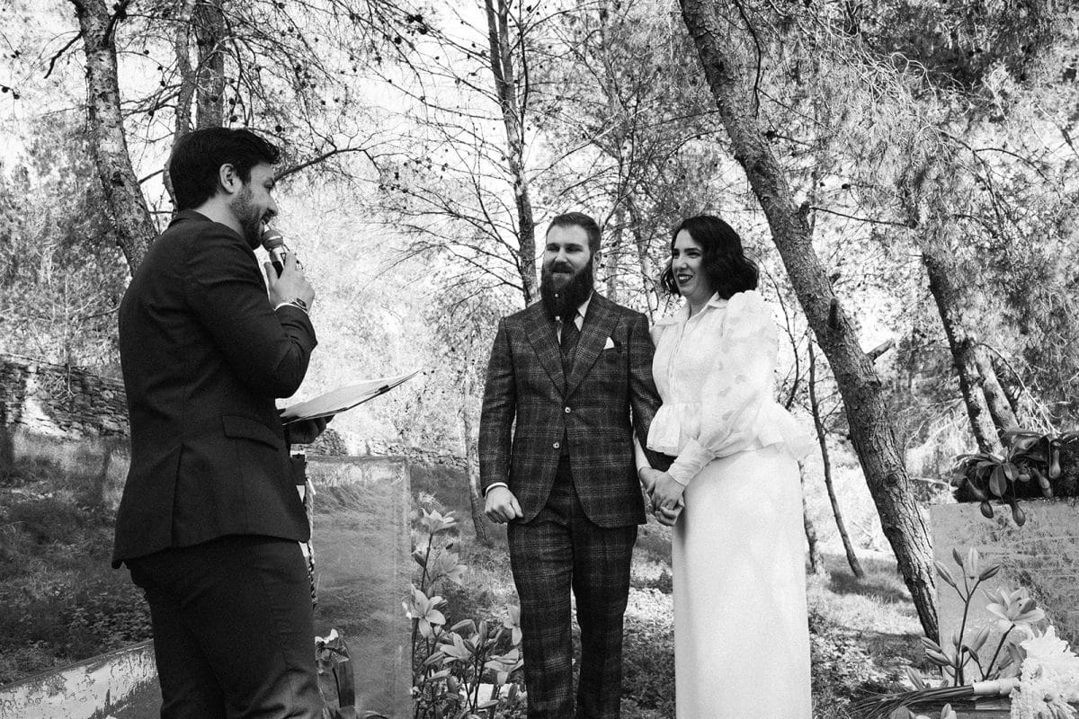 Pareja de novios en ceremonia civil al aire libre con sillas madera, flores frescas y decoración natural entre árboles.